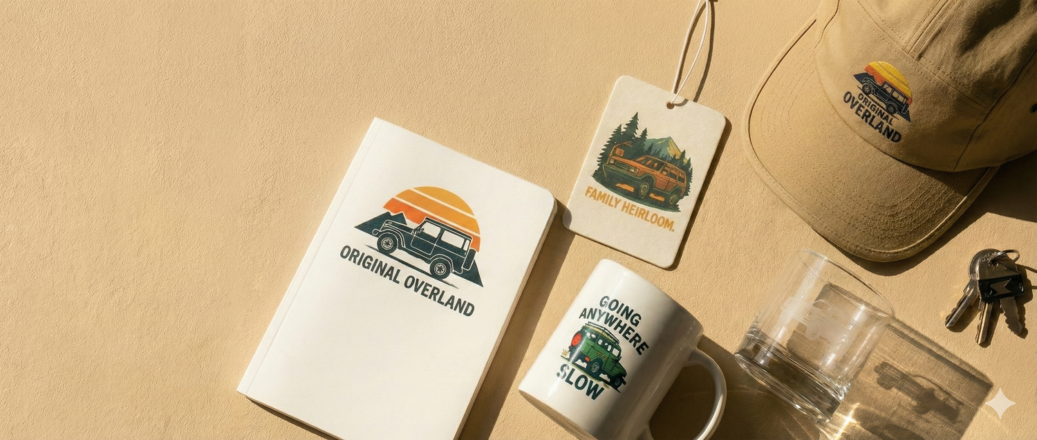 A top-down flat lay photograph for the homepage hero slider, featuring Original Overland accessories like a vintage Toyota Land Cruiser coffee mug, tan 5-panel hat, notebook, and air freshener arranged on a warm beige background with space on the left.