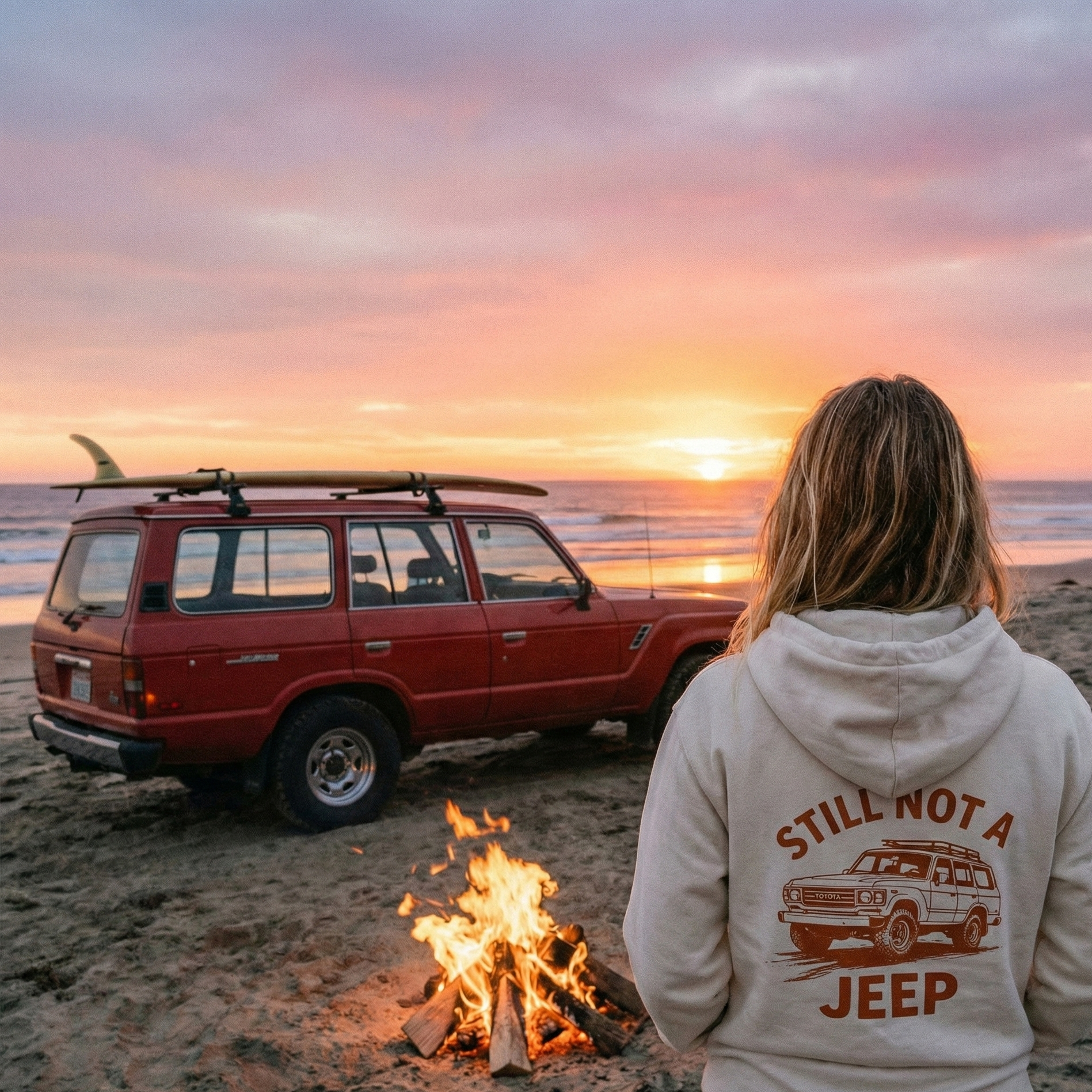A candid shot from behind of a woman standing on a sandy beach at sunset, wearing the off-white 'STILL NOT A JEEP' hoodie with the Toyota Land Cruiser graphic. She is next to a bonfire, looking at a red FJ60 Land Cruiser parked on the sand with a surfboard on its roof rack.